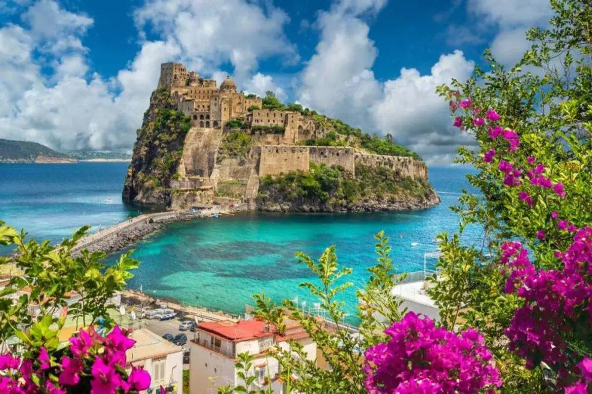 Un castello su uno scoglio domina un mare cristallino. La vittoria colonna di questo luogo incantevole &egrave; esaltata dai fiori fucsia in primo piano.