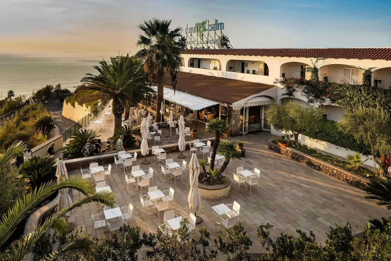 Terrazza con tavoli e sedie all'aperto del hotel Le Palme Ischia, con vista sul mare al tramonto e palme rigogliose.