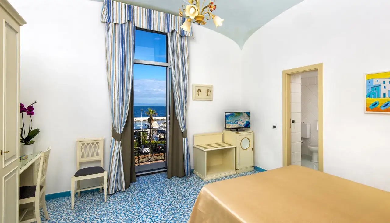 Camera con vista mare all'Hotel Svizzera Ischia. Pavimento decorato, tende a righe e un quadro di barche blu.