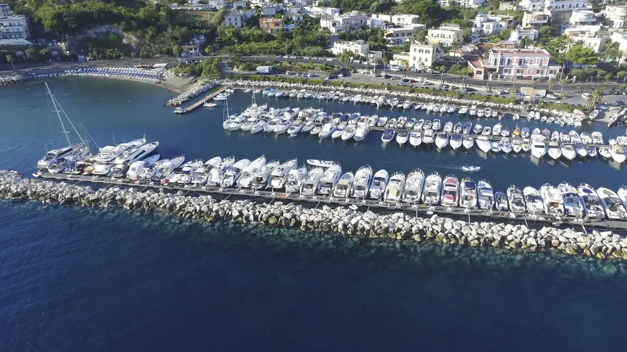 Un affascinante ormeggio a Ischia, con decine di yacht e barche ormeggiate lungo un molo di roccia, sullo sfondo di una pittoresca cittadina costiera.