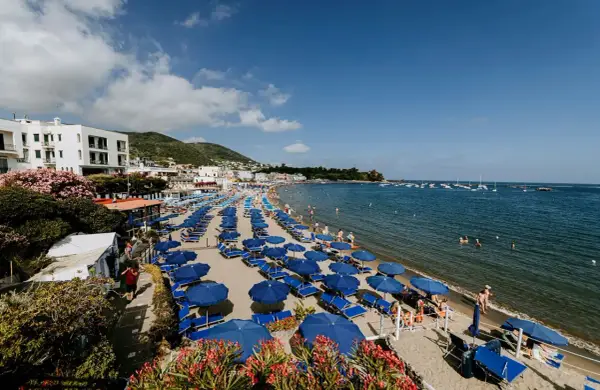 Spiaggia affollata a Ischia Lido con file di ombrelloni blu e lettini, bagnanti in acqua e barche all'orizzonte.