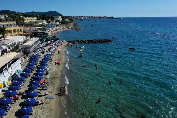 Forio Ischia spiagge affollate sotto il sole. Ombrelloni blu, bagnanti in mare e hotel affacciati sulla costa.