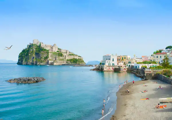 Lidi di Ischia Porto con il Castello Aragonese sullo sfondo. Gente in spiaggia e mare azzurro sotto un cielo limpido.